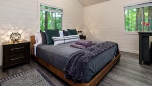 Ein Schlafzimmer mit schrägem Dach, einem Oberlicht, einem Bett mit Holzrahmen, einem Nachttisch, einem Schreibtisch und einem Fenster mit Blick auf Bäume.