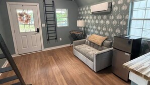 Un espai tipus loft amb sòl de fusta, un sofà gris, una taula petita, un frigorífic i una finestra amb vistes.