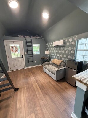 A loft-style room with a wooden floor, a gray sofa, a small table, a refrigerator, and a window with a view.