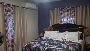 A bedroom with a bed, a bench, curtains, an air conditioner, and a fan.