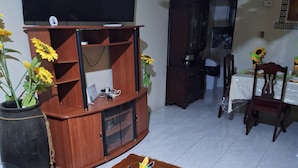 A living room with a wooden entertainment center, a coffee table with decorative items, a TV mounted on the wall, and a dining area with a table and chairs.