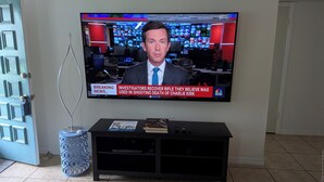 A television mounted on a wall displaying a news broadcast. - Amazing move in ready condo just 10 minutes from the beach. 65 plus community. (St Petersburg)