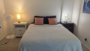 A neatly made bed with striped bedding, two pillows, a bedside table with a lamp, a floor fan, and a wall-mounted light fixture. - Amazing move in ready condo just 10 minutes from the beach. 65 plus community. (St Petersburg)