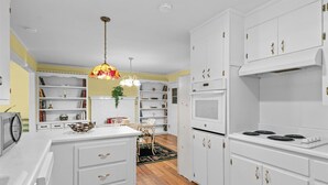 Une cuisine moderne avec des meubles blancs, un four encastré et un four à micro-ondes. On y trouve un coin repas avec une table et des chaises blanches, et un luminaire suspendu au-dessus.