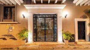 Entrada de hotel com um portão preto ornamentado, plantas em vasos e uma placa com o número 10-05.
