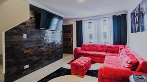 A living room with a red sectional sofa, a red ottoman, a black and white rug, a stone wall, a flat-screen TV, and a window with curtains.