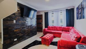 Una sala de estar con un sofá rojo, un televisor negro montado en una pared de piedra, y una ventana con cortinas.