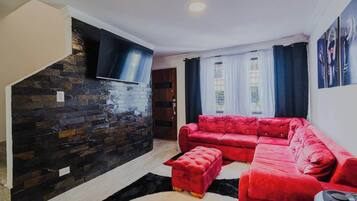 A living room with a red sofa, a black TV mounted on a stone wall, and a window with curtains.