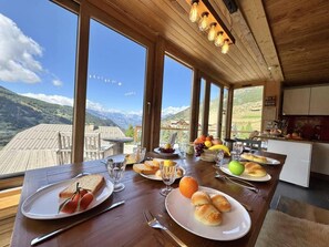 Une table à manger dressée avec des aliments pour le petit-déjeuner, de grandes fenêtres offrant une vue sur les montagnes et un plafond en bois agrémenté de luminaires suspendus.