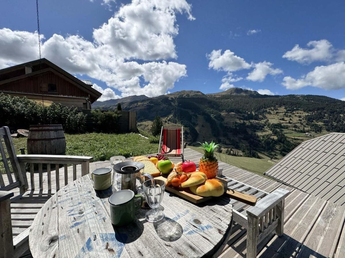 A wooden deck with a round table set for a picnic, featuring fruits, bread, and drinks, overlooking a mountainous landscape.