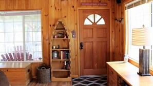 Interior de una cabaña de madera con una puerta de madera, una estantería, una ventana que muestra sillas al aire libre y una lámpara sobre una mesa de madera.