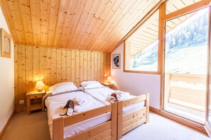 A wooden cabin bedroom with a bed, two bedside tables, and a large window offering a view of a snowy landscape.