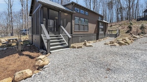 A modern two-story cabin with a covered porch, a staircase leading to the entrance, and a gravel driveway.