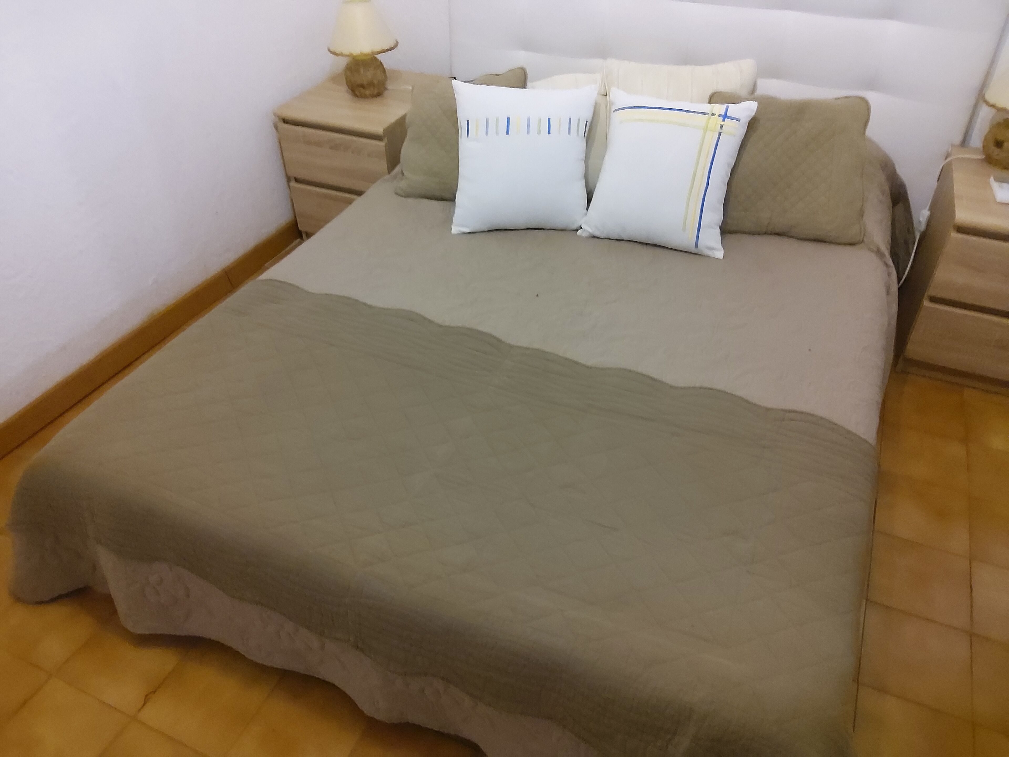 Een netjes opgemaakt bed met een beige deken en kussens, een bedlamp en een houten nachtkastje.