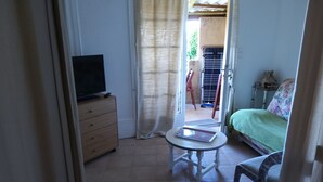 A room with a television on a wooden dresser, a small round table with a notebook, a bed with a floral bedspread, and a view of an outdoor area with a chair and a table. - Seaside vacations (Coti-Chiavari)