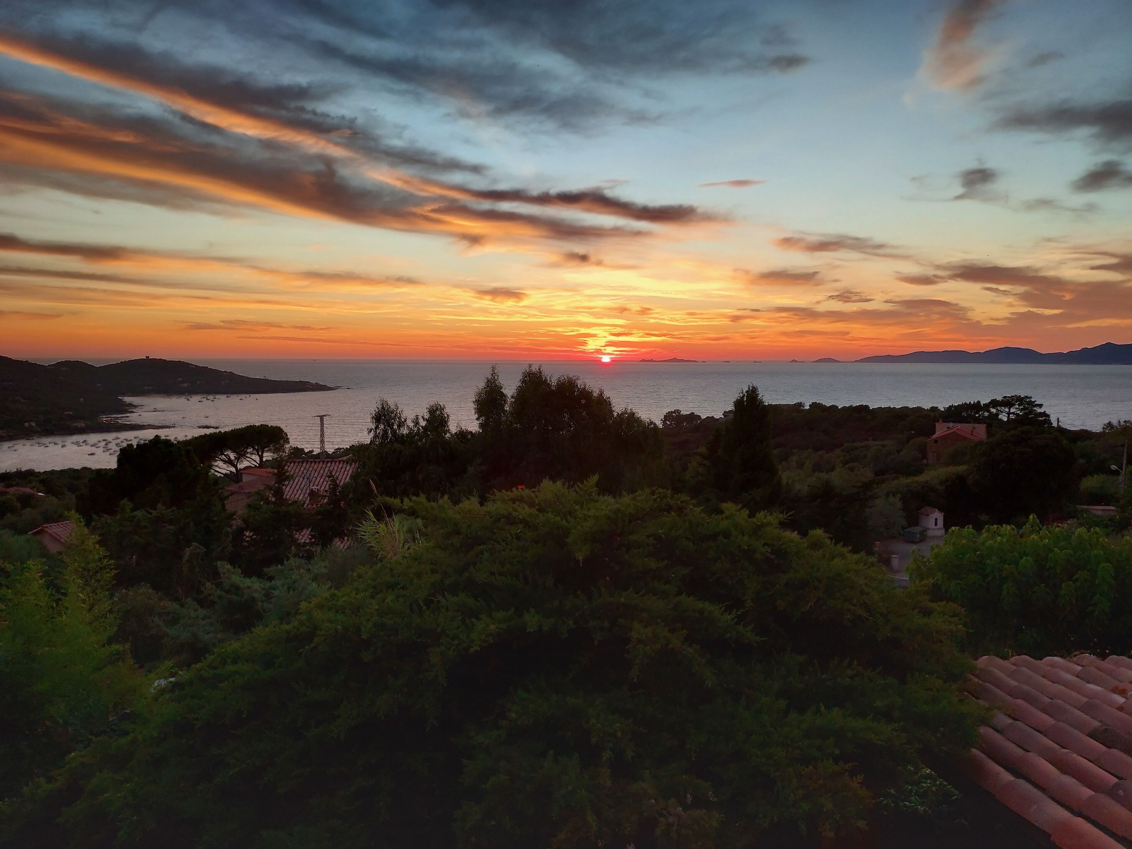 A coastal landscape with a sunset over the sea, hills in the background, and a view of the sea with boats.