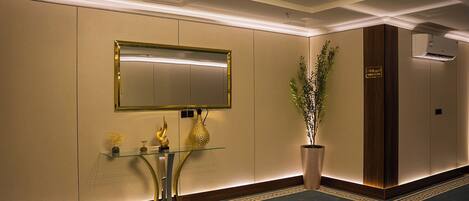 A modern hotel lobby with a large mirror, a reception desk, a potted plant, and a wall-mounted air conditioning unit.