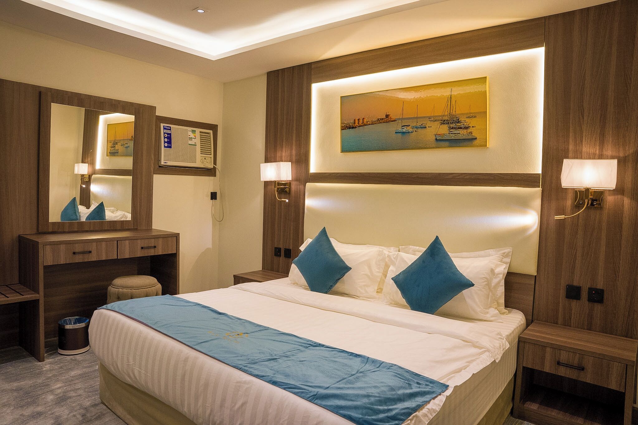 Una habitación de hotel con una cama grande, un cabecero de madera, dos mesitas de noche con lámparas, un espejo y un cuadro de un puerto.