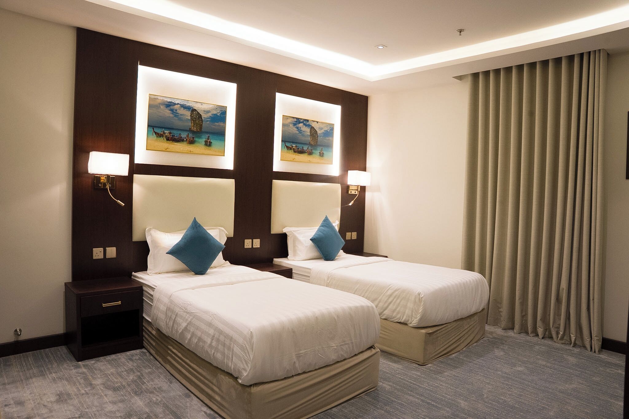Habitación de hotel con dos camas, cada una con cabecero, mesitas de noche y lámparas. Hay dos cuadros en la pared sobre las camas.