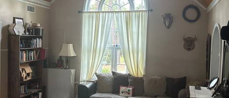 Ein gemütliches Wohnzimmer mit hoher Holzbalkendecke, einem großen Bogenfenster, einem Bücherregal, einer Couch, einem Couchtisch und einem Spiegel.