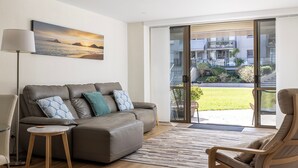 Ein modernes Wohnzimmer mit einer Couch, einem kleinen Tisch und einem Sessel. Ein großes Fenster bietet Blick auf ein Gebäude und eine Rasenfläche.