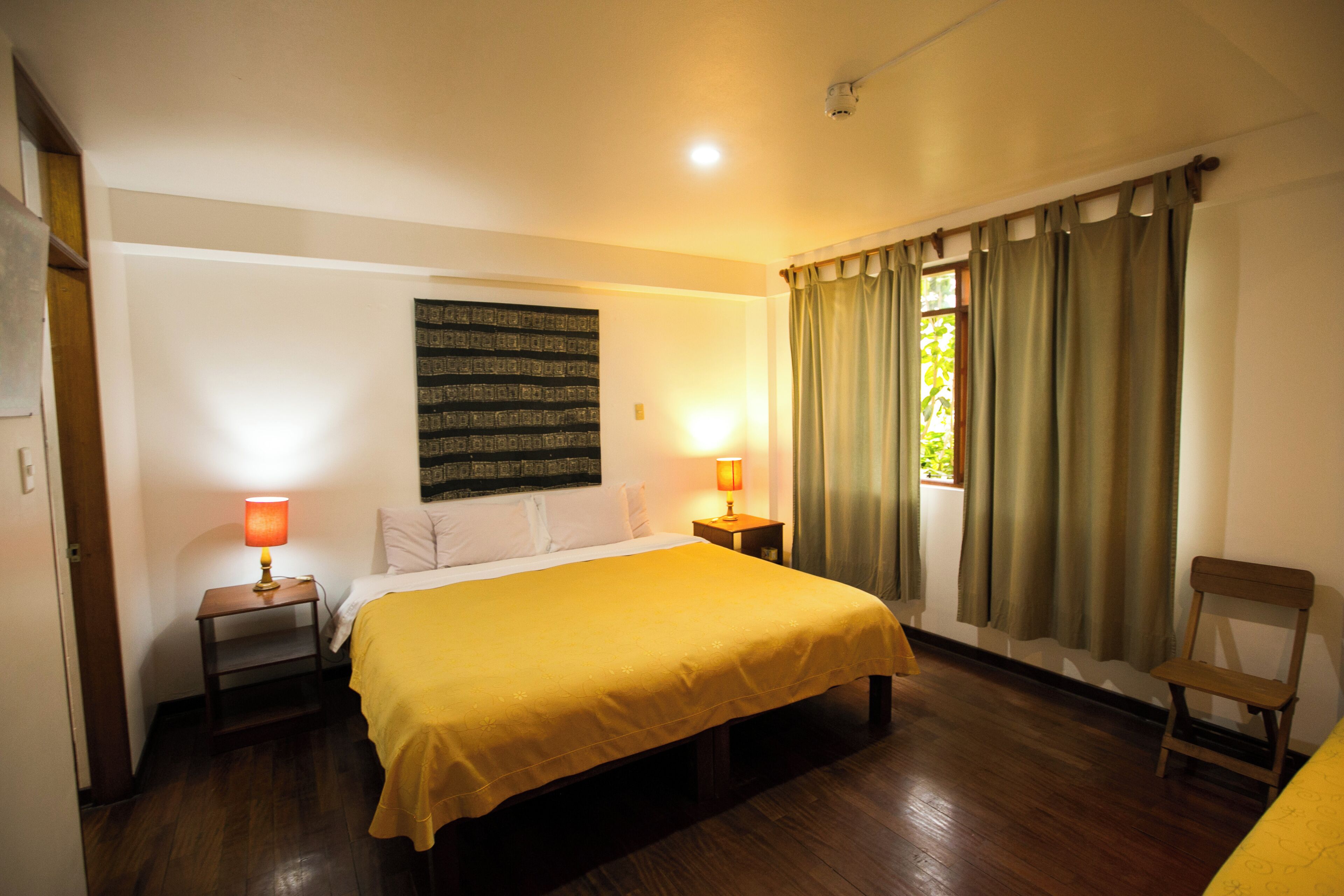 A hotel room with a bed, two bedside lamps, a window with curtains, a wooden floor, and a wall-mounted artwork.