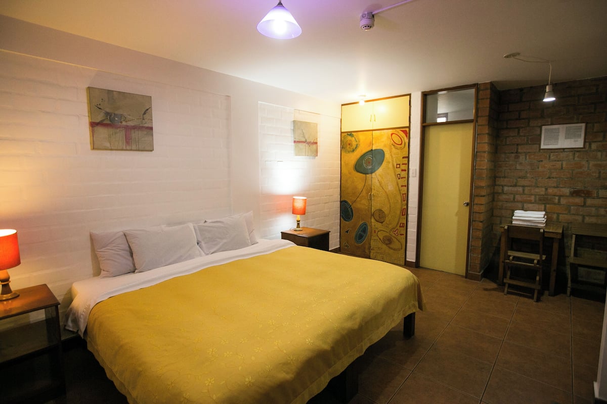 A hotel room with a bed, two bedside lamps, a small table with a lamp, a brick wall, and a door.