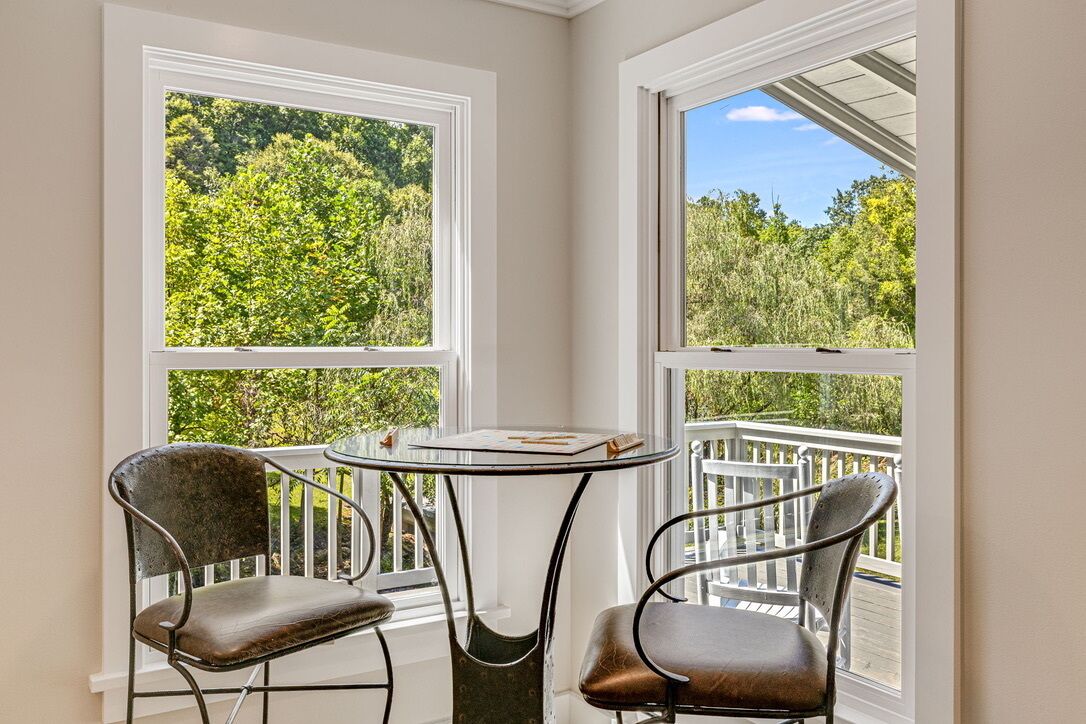 A corner room with two chairs and a small table, large windows with a view of greenery, and a balcony with a railing.
