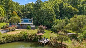 Une maison à deux étages avec un porche, entourée d’une végétation luxuriante et bordée d’un étang.