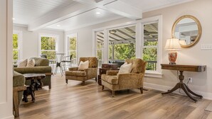 Un vaste salon doté d’un plafond haut, de grandes fenêtres et offrant une vue sur la verdure environnante. On y trouve plusieurs canapés et fauteuils disposés autour d’une table basse.