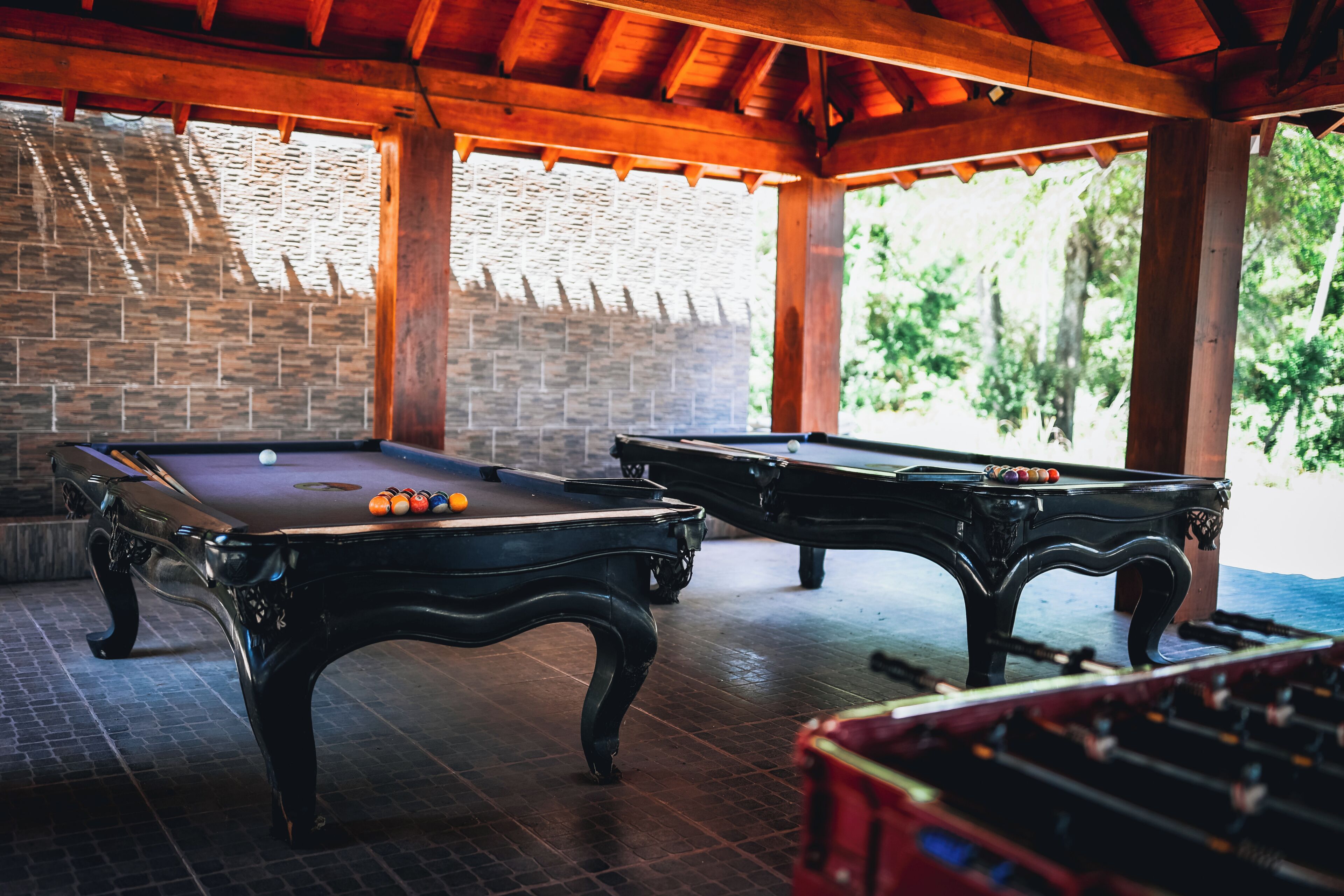 Two pool tables in a covered area with a brick wall and wooden beams.