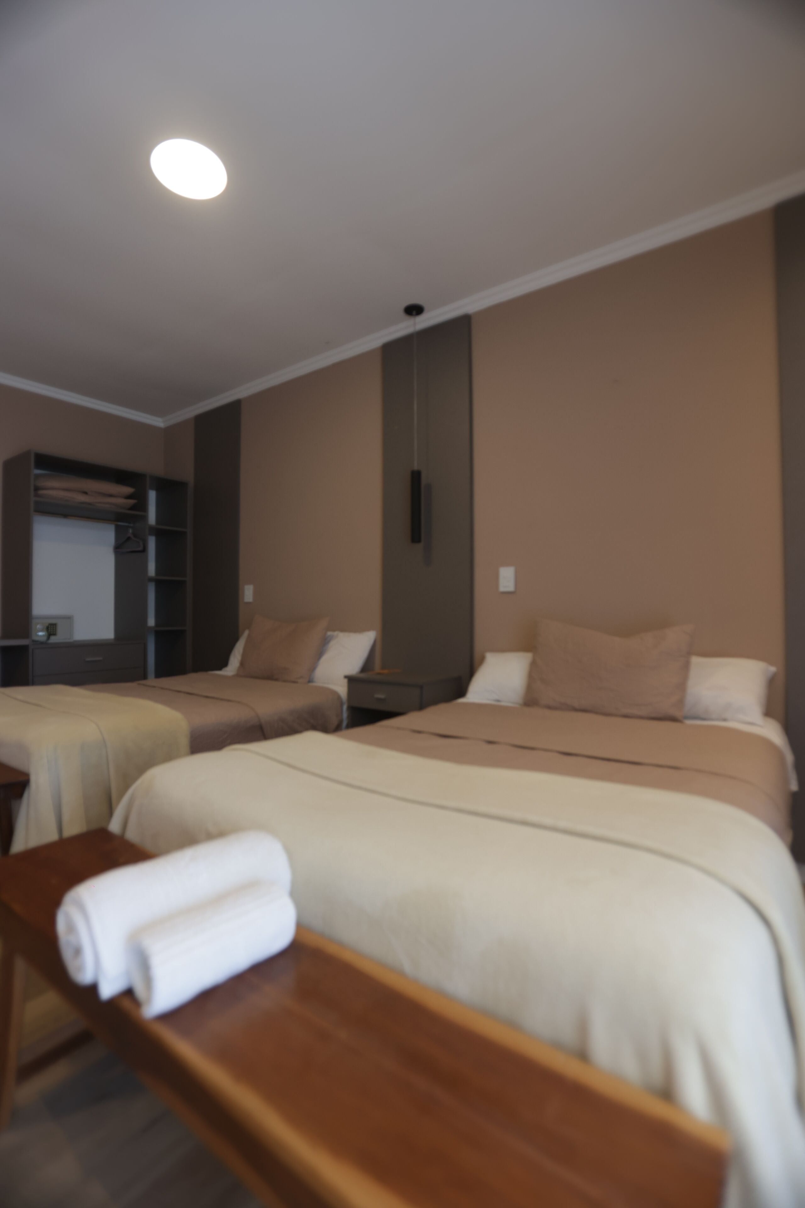 A hotel room with two beds, a wooden bench, a wardrobe, and a ceiling light.