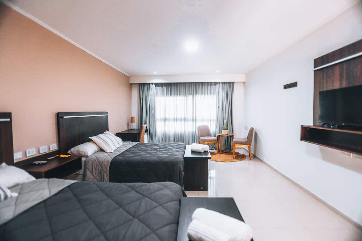 Habitación de hotel con dos camas, un escritorio, una silla, un televisor montado en la pared y una ventana con cortinas.
