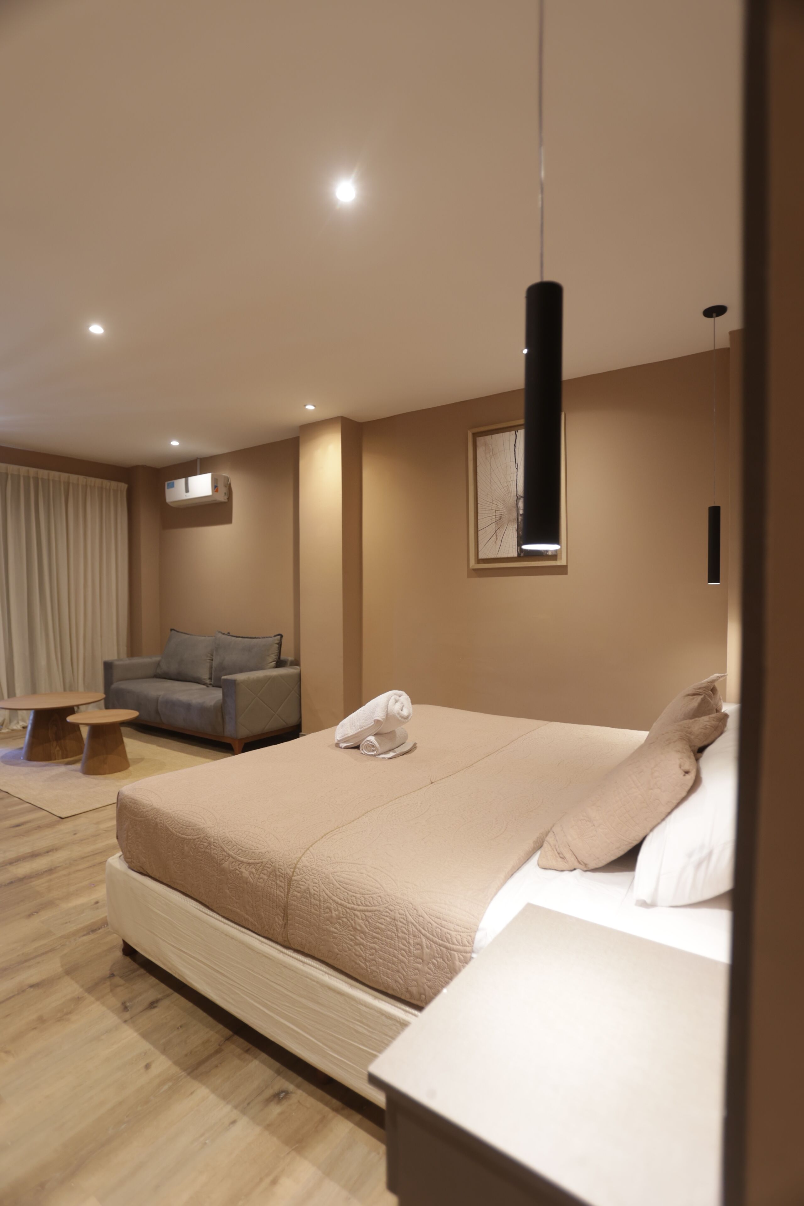 A modern hotel room with a large bed, a sofa, a small table, and a hanging light fixture.