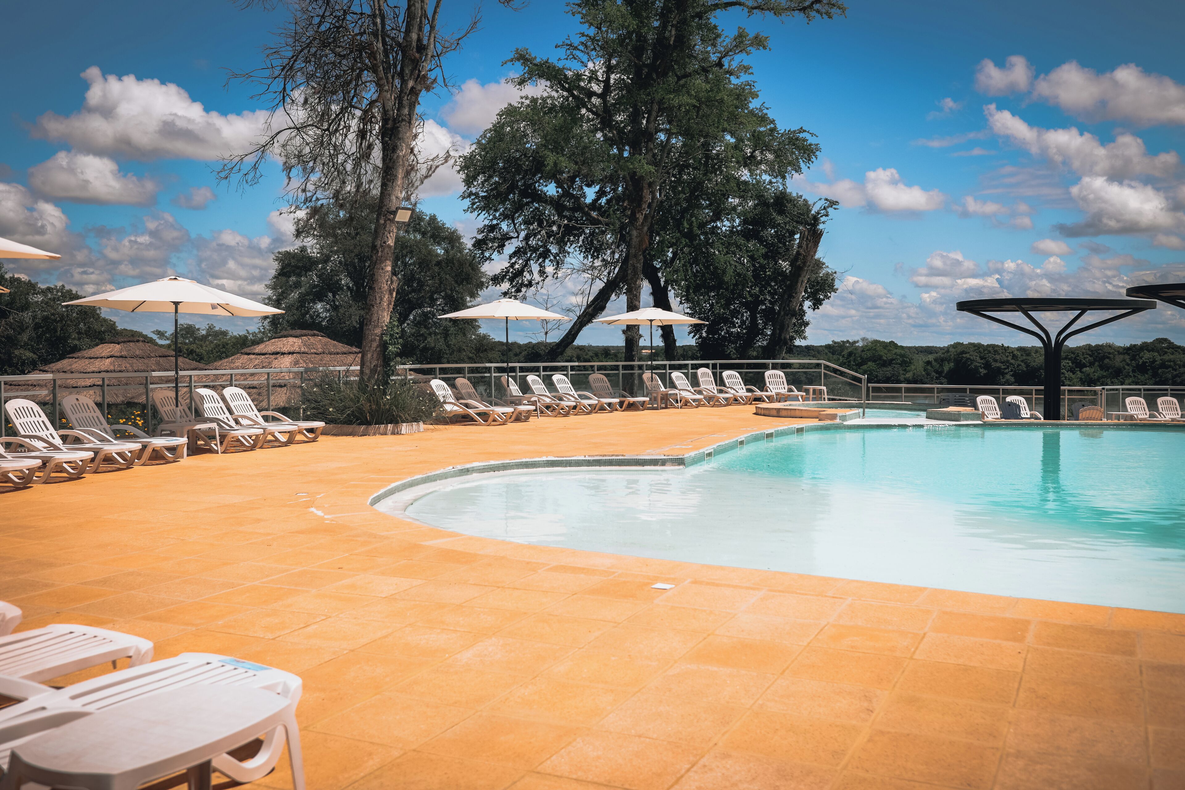 A pool area with lounge chairs and umbrellas, surrounded by trees and a clear sky.