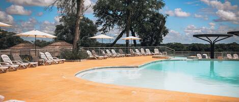A pool area with lounge chairs and umbrellas, surrounded by trees and a clear sky.