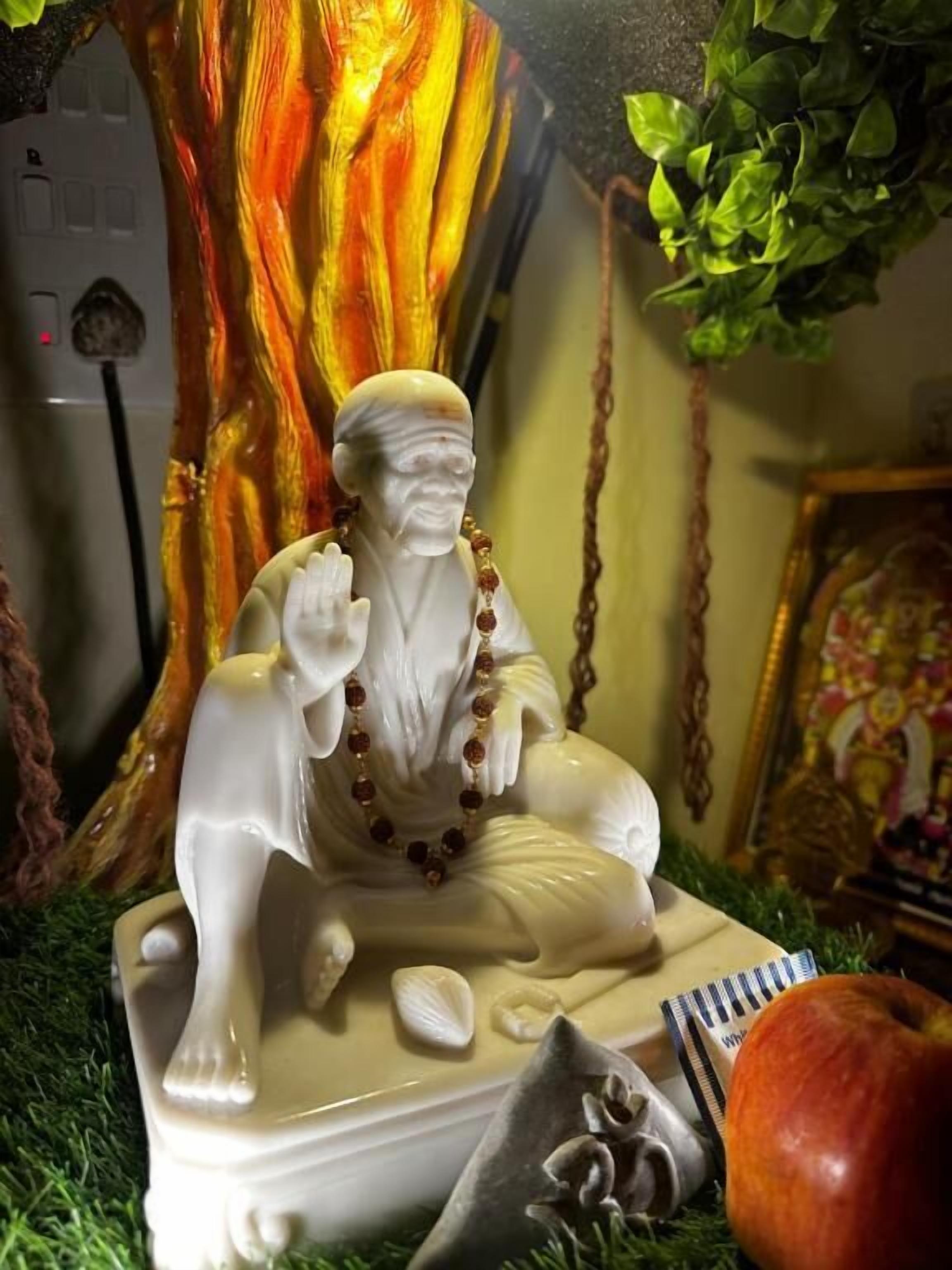A statue of a seated figure with a white dog, surrounded by decorative elements like seashells and a book, set against a backdrop of a tree and a framed picture.