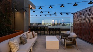 Ein Außenbereich mit Sitzgelegenheiten, darunter eine Couch, Stühle und ein Tisch, eine Bar mit Hockern sowie ein Blick auf die Skyline der Stadt.