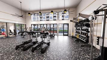 A modern gym with treadmills, stationary bikes, and weight machines. It features large windows, ceiling fans, and a weight rack.
