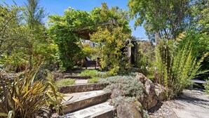 A garden with stone steps leading to a wooden structure, surrounded by various plants and trees. - Birdsong House, a contemporary family home close to the beach. (Kaiteriteri)