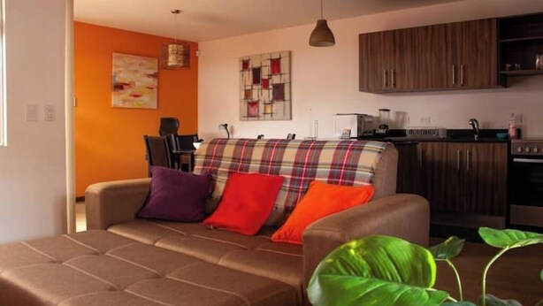 A living room with a sofa, a coffee table, and a kitchen area with wooden cabinets and a countertop. - Sabana Sur House (San José)