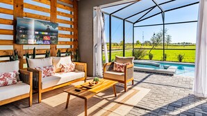 Un espacio exterior moderno con techo de madera, una gran pantalla de televisión y vistas a una piscina y campos verdes.