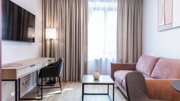 A modern hotel room with a wooden desk, black chairs, a small table with a vase, a pink sofa, and a large window with curtains.