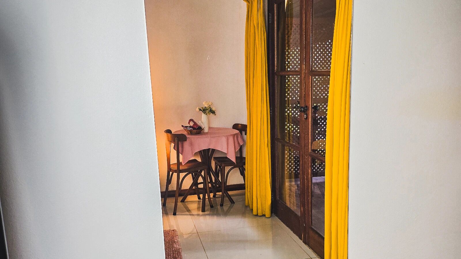 Uma sala de jantar com uma mesa posta para dois, uma cadeira e uma pequena mesa com um vaso e um livro.