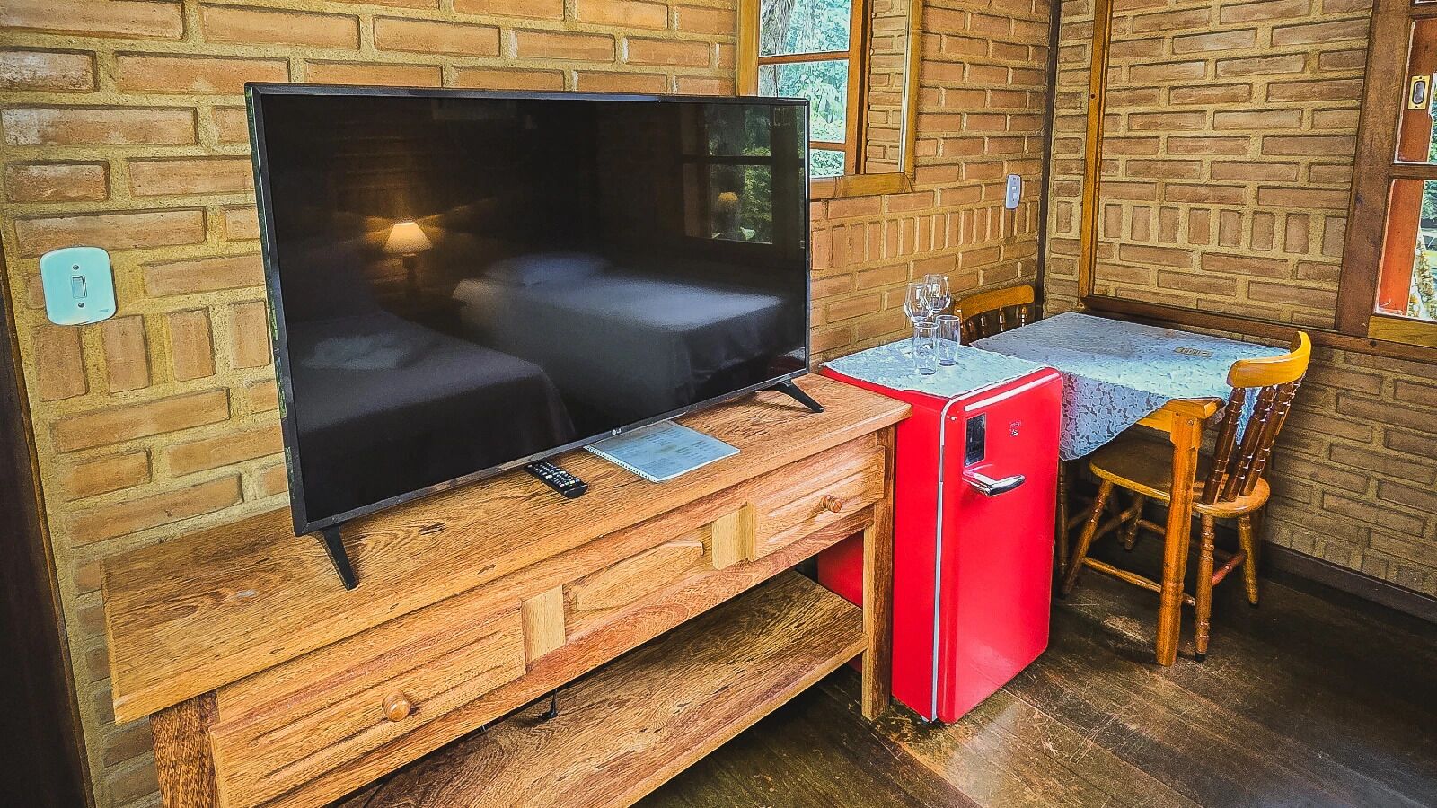 A cozy room with a TV on a wooden stand, a small red fridge, a wooden table with chairs, and a brick wall.