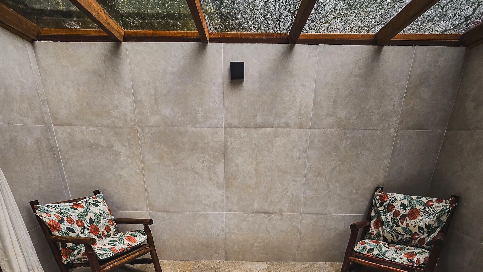A room with two wooden chairs with floral upholstery, a tiled wall, and a small black object on the wall.