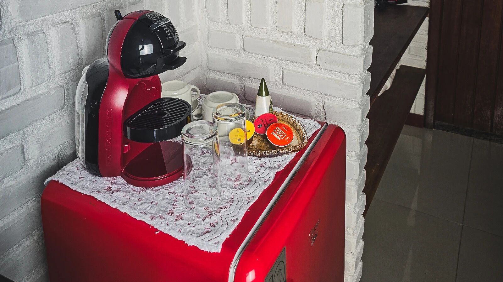 Un electrodoméstico rojo sobre un mantel de encaje blanco, con diversos utensilios de cocina y un frasco de vidrio.