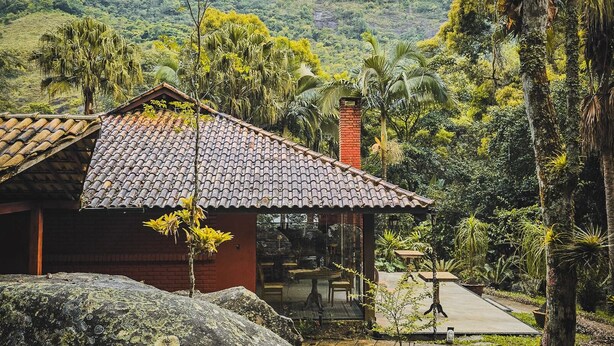 Una casa rústica con techo de tejas, una chimenea de ladrillo y un ventanal que da a un espacio de estar al aire libre rodeado de exuberante vegetación.