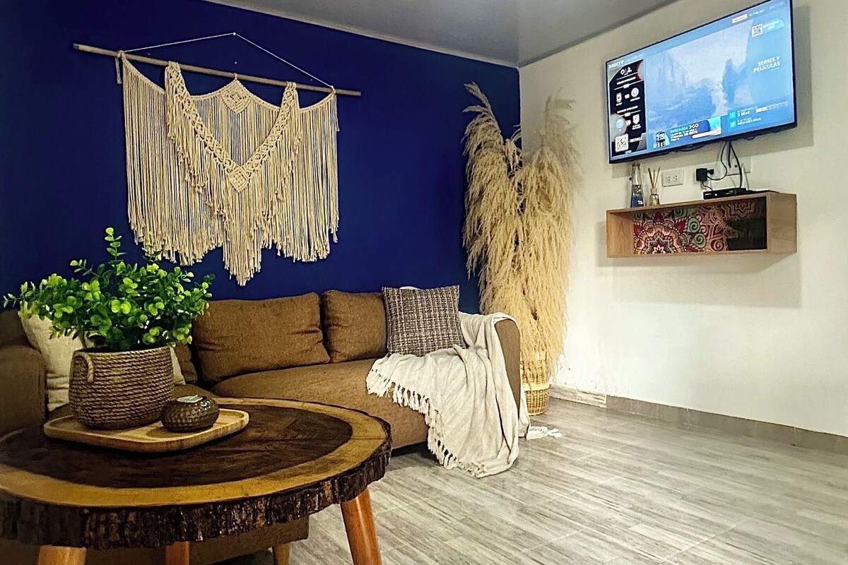 Un salon avec un mur d’accent bleu, une table basse ronde en bois, un canapé marron, une décoration murale en macramé, une grande plante et un téléviseur fixé au mur.