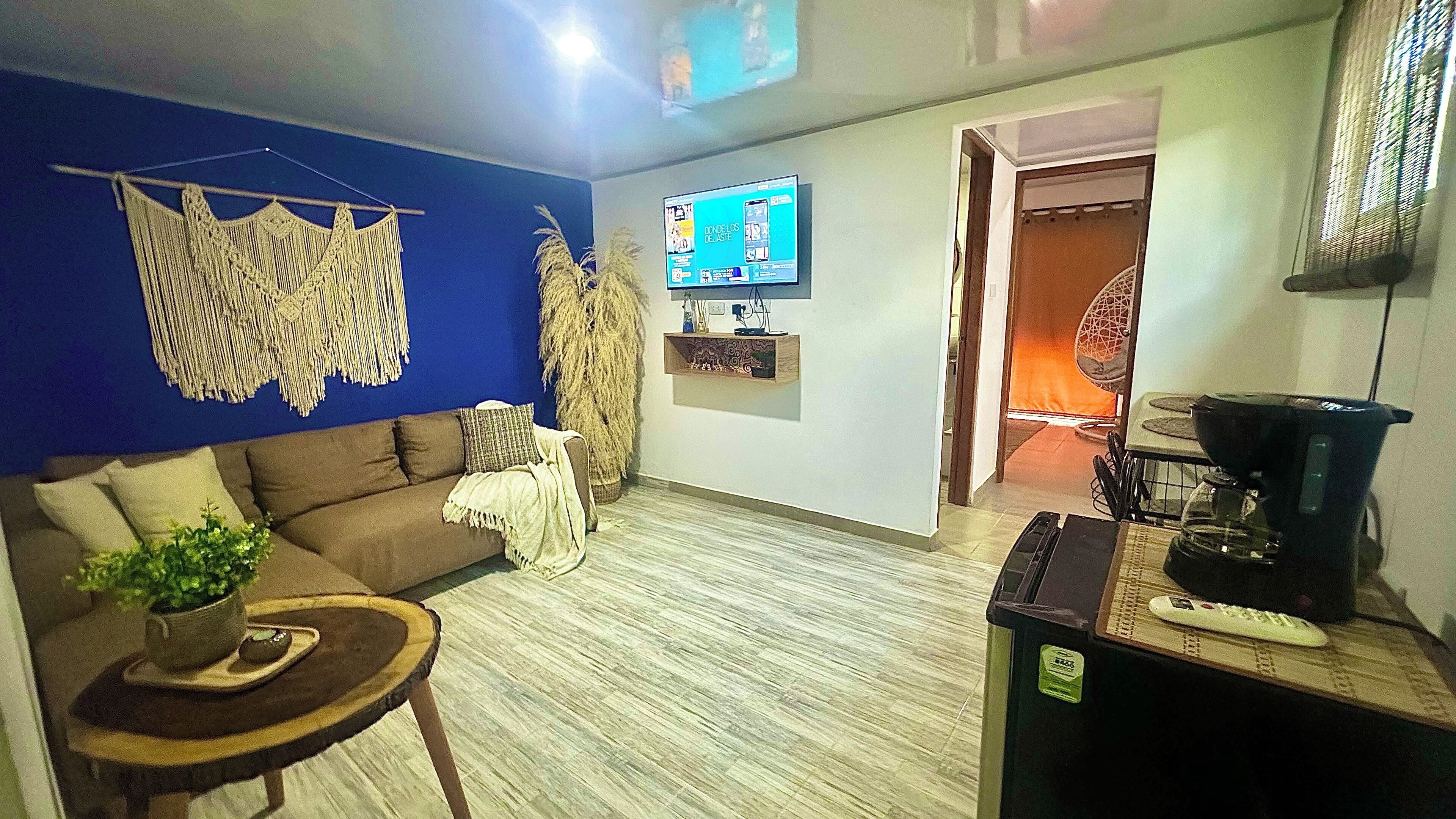 A living room with a sofa, a coffee table, a TV, and a kitchen area with a blender.